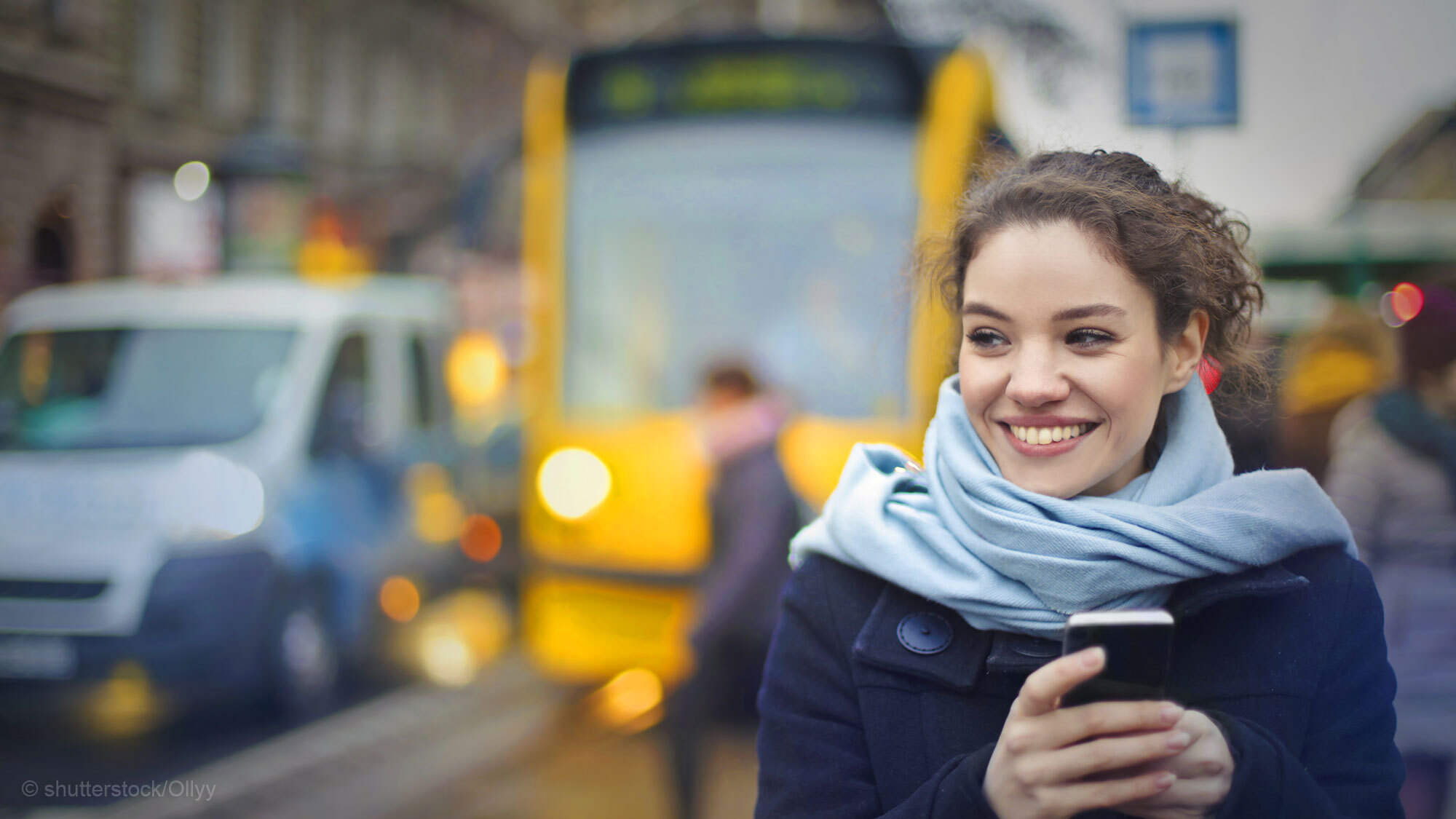 Frau steht mit Handy in der Hand an der Straßenbahnhaltestelle