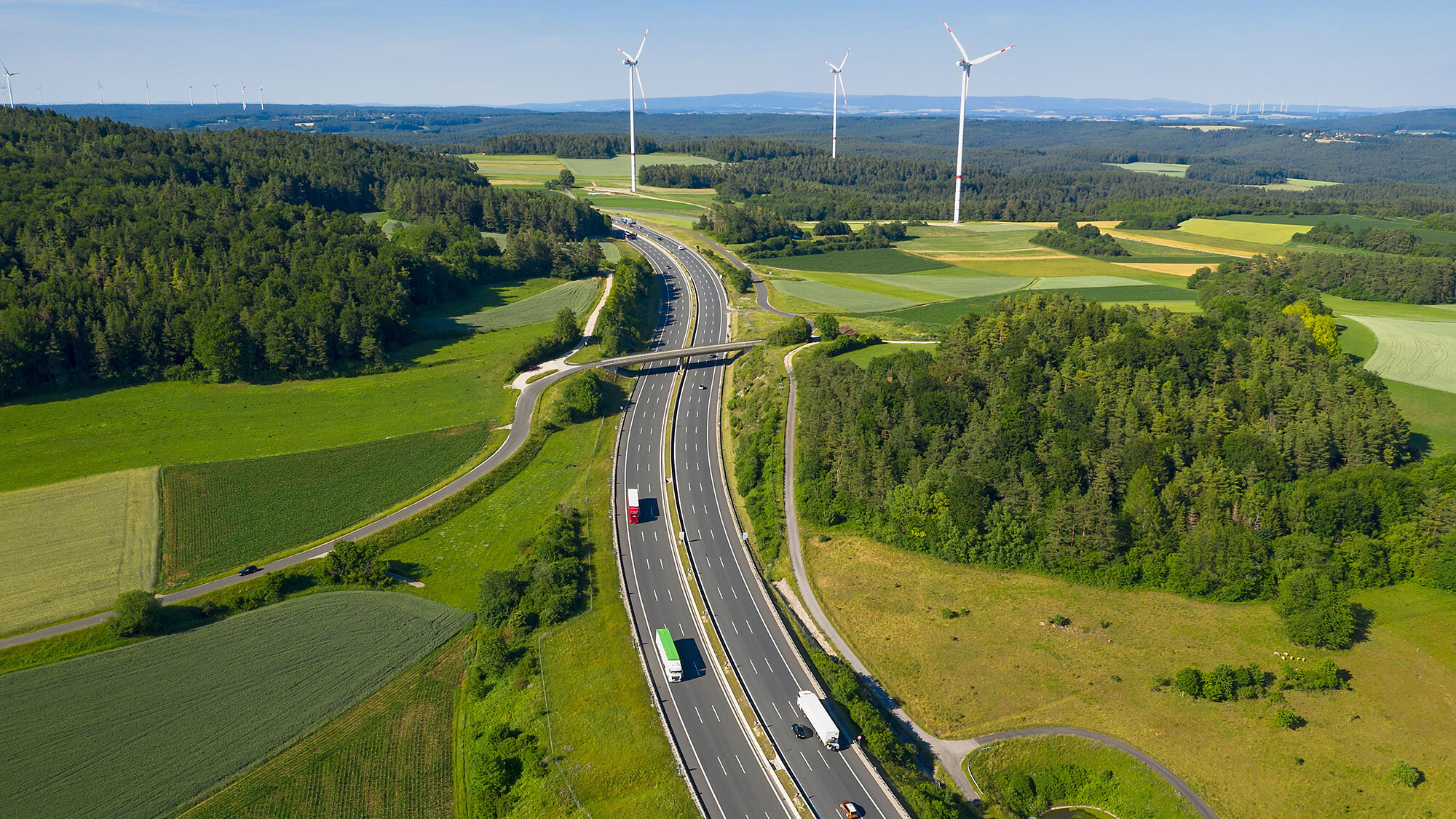 Autobahn führt durch Natur, grüne Wiesen, Windräder am Horizont, auf der Autobahn fahren Pkw und LKW. Text auf Bild: Mehr Tempo bei Straßen, mehr Marktwirtschaft beim Klimaschutz