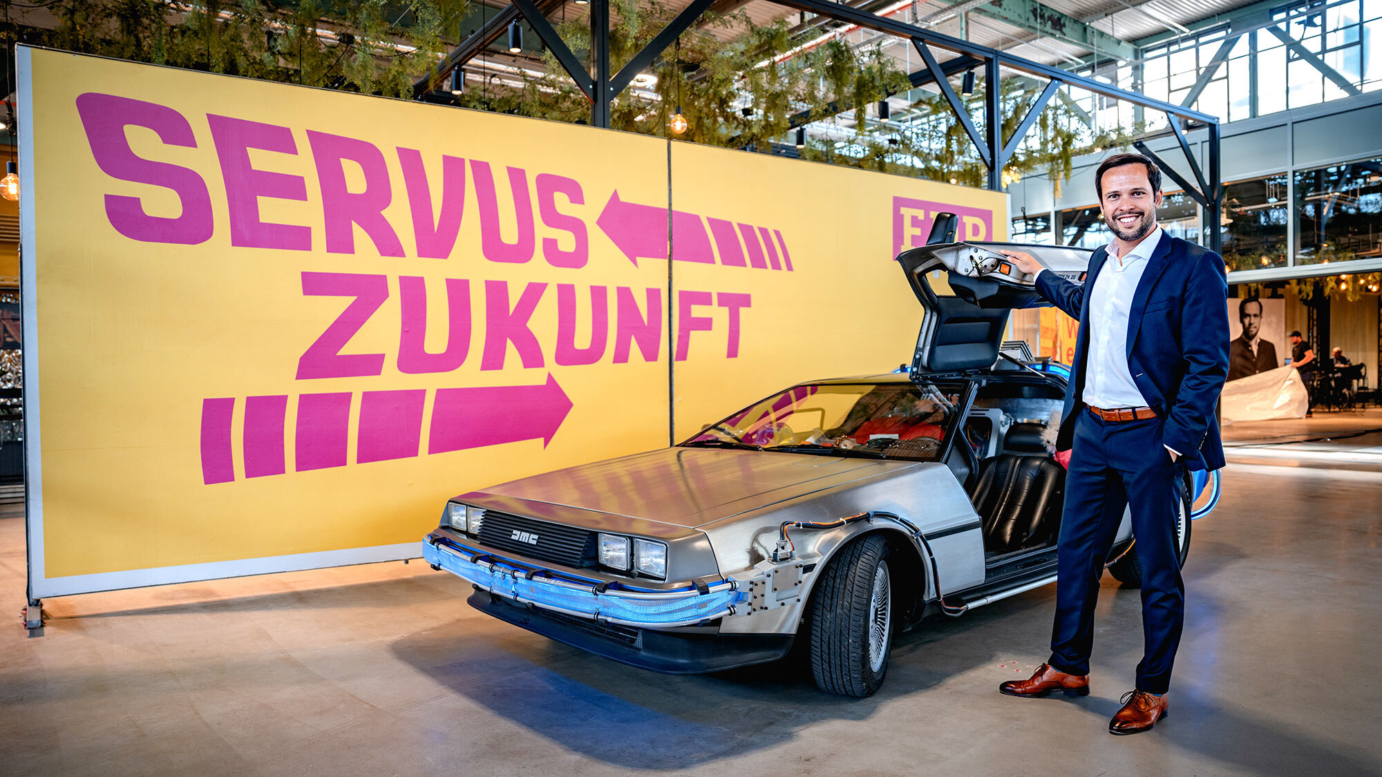 Spitzenkandidat der FDP Bayern, Martin Hagen, steht vor einem DeLorean DMC-12 vor einem großen Plakat, auf dem groß "Servus Zukunft" steht.