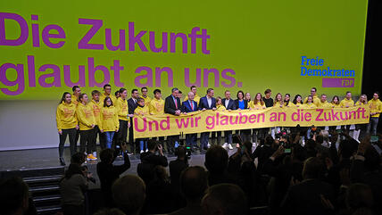 Bühne beim Dreikönigstreffen der Freien Demokraten, im Hintergrund steht "Die Zukunft glaubt an uns." und auf dem Banner, das von jungen Menschen und FDP-Politikerinnen und -Politikern hochgehalten wird, steht: "Und wir glauben an die Zukunft."