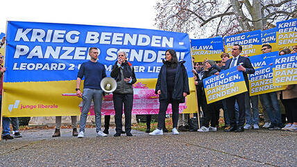 Marie-Agnes Strack-Zimmermann und Franziska Brandmann stehen vor einem Transparent vor dem Dreikönigstreffen, auf dem steht: Krieg beenden, Panzer senden! Eine Aktion der Jungen Liberalen