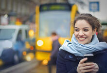 Frau steht mit Handy in der Hand an der Straßenbahnhaltestelle