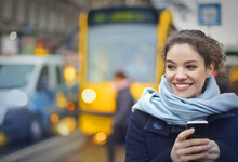 Frau steht mit Handy in der Hand an der Straßenbahnhaltestelle