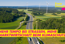 Autobahn führt durch Natur, grüne Wiesen, Windräder am Horizont, auf der Autobahn fahren Pkw und LKW. Text auf Bild: Mehr Tempo bei Straßen, mehr Marktwirtschaft beim Klimaschutz