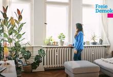 Frau, die in einem Wohnzimmer am Fenster an den Heizkörpern steht und nach draußen schaut.