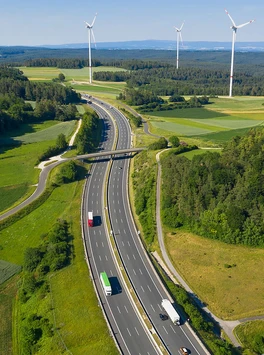 Autobahn führt durch Natur, grüne Wiesen, Windräder am Horizont, auf der Autobahn fahren Pkw und LKW. Text auf Bild: Mehr Tempo bei Straßen, mehr Marktwirtschaft beim Klimaschutz