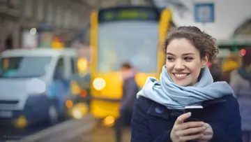 Frau steht mit Handy in der Hand an der Straßenbahnhaltestelle