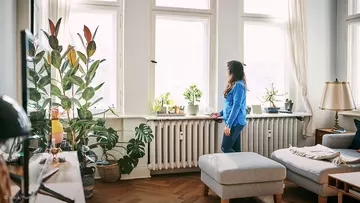 Frau steht in Wohnung am Fenster vor zwei Heizkörpern
